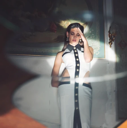 Woman in a white sleeveless dress with black trim posing indoors with a painting and warm spotlight.