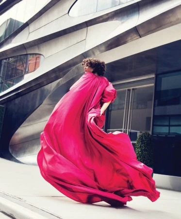 Model in a flowing magenta gown posing outdoors in front of a futuristic metallic building exterior.