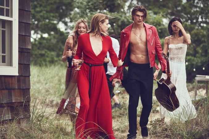 Group of models walking outdoors through tall grass with a red dress, red leather jacket, and holding a guitar and champagne glass.