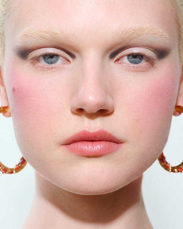 Close-up beauty portrait of a model with bleached brows, dramatic dark eyeshadow, flushed pink cheeks, and gold hoop earrings against white