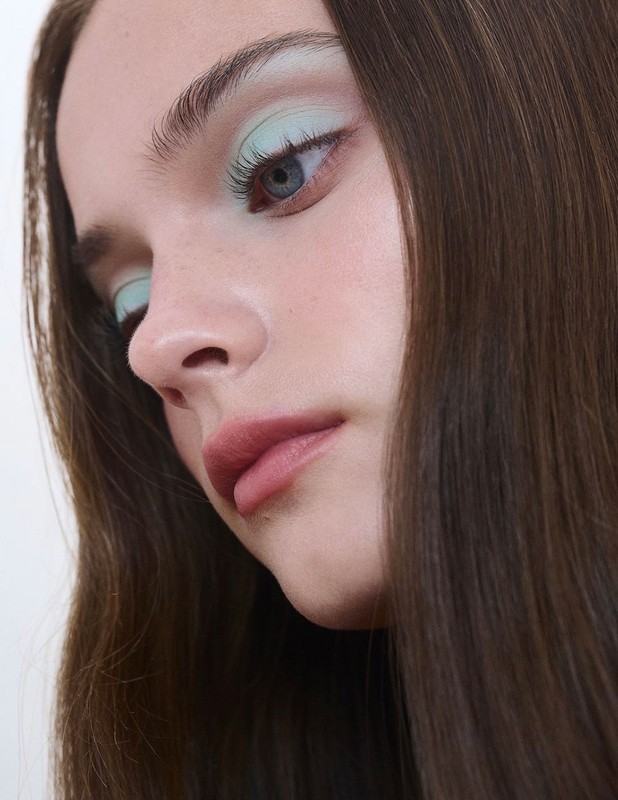 Portrait of a young woman with long brown hair, light skin, and blue eye wearing light blue eyeshadow, in an indoor setting.