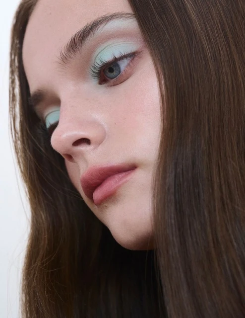 Portrait of a young woman with long brown hair, light skin, and blue eye wearing light blue eyeshadow, in an indoor setting.
