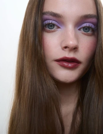 Close-up portrait of a woman with long brown hair, purple eyeshadow, and dark glossy lips against a white background.