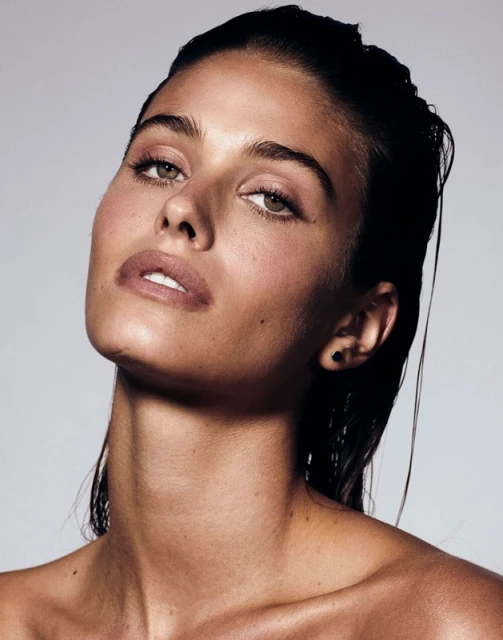 Close-up portrait of a young, tan-skinned woman with wet, dark hair, light eyes, and nude makeup, gazing upwards against a grey background.