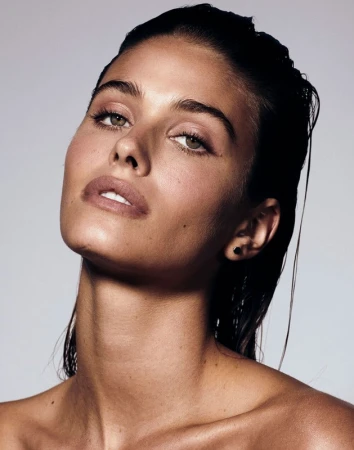 Close-up portrait of a young, tan-skinned woman with wet, dark hair, light eyes, and nude makeup, gazing upwards against a grey background.
