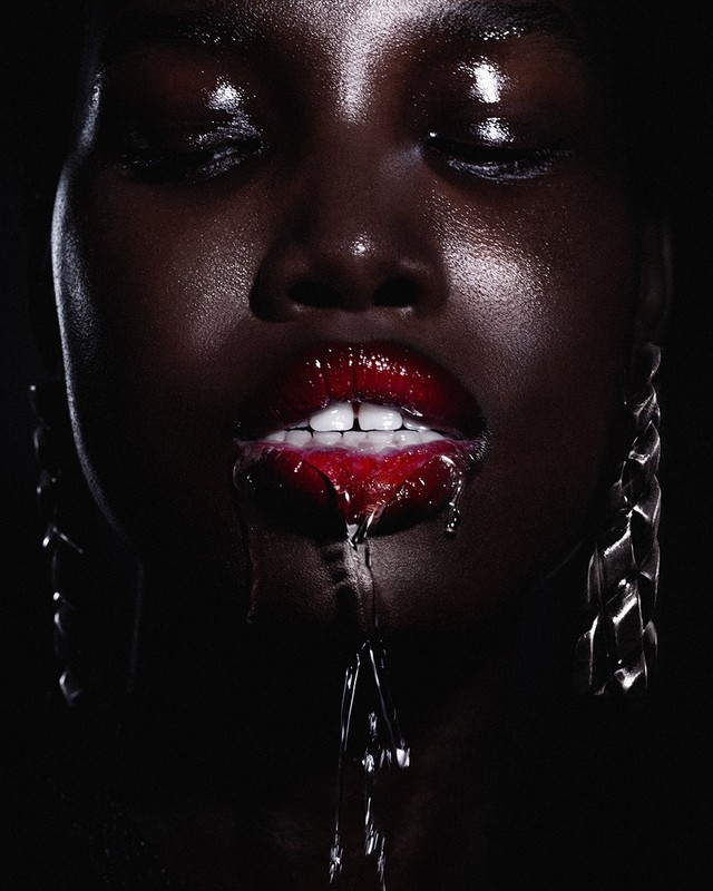 Close-up of an African American woman with dark skin, glossy red lips with liquid dripping, closed eyes, and silver chain earrings.