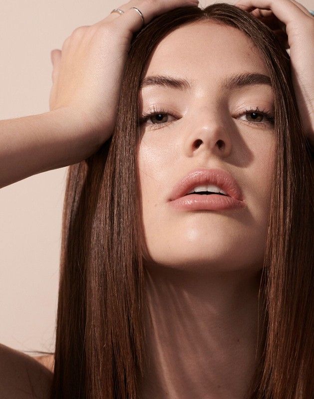Close-up beauty portrait of a woman with long brown hair framing her face, hands resting gently on her head against a neutral background.