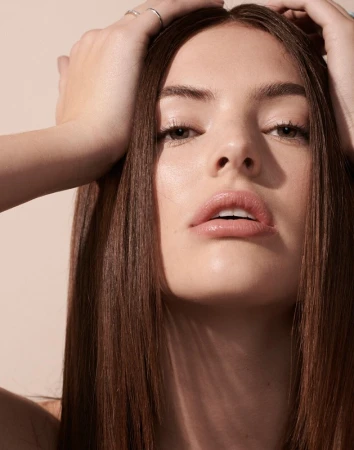Close-up beauty portrait of a woman with long brown hair framing her face, hands resting gently on her head against a neutral background.
