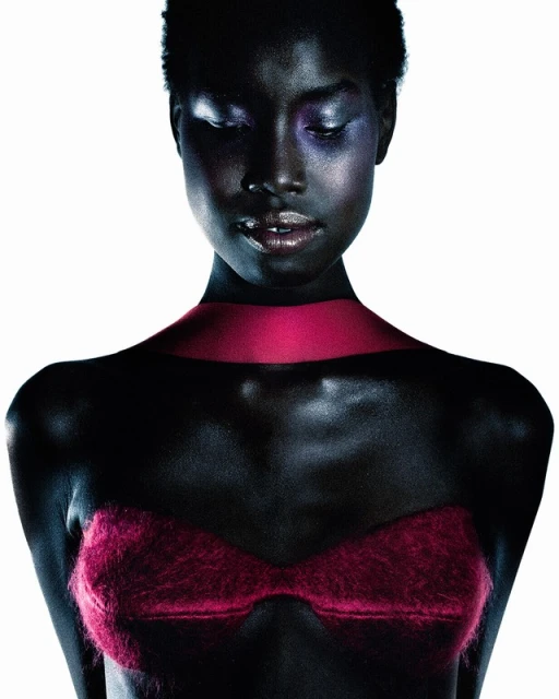 Dark-skinned woman with colorful eyeshadow, wearing a pink bra and collar, poses in a studio setting with a white background.