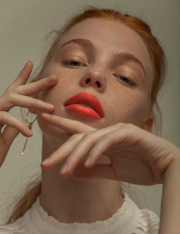 Close-up portrait of a red-haired woman with bright red lipstick posing indoors with her hands near her face, gazing at the camera.