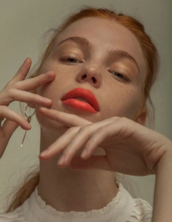 Close-up portrait of a woman with red hair and bright coral lipstick holding her hands near her face
