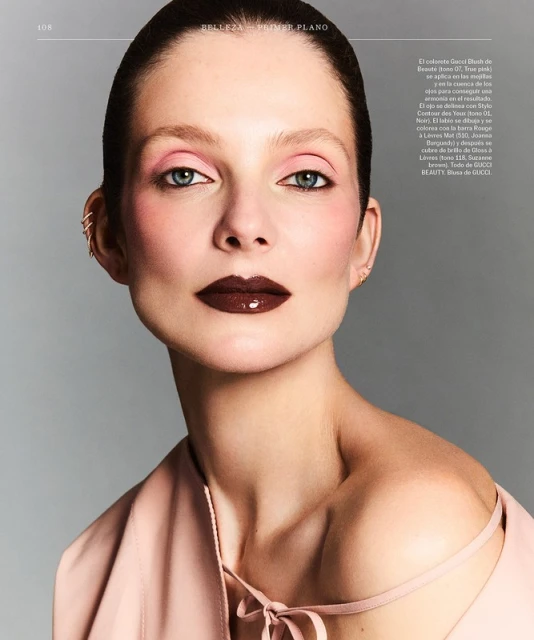Beauty shot of a woman with pink eye makeup and dark glossy lipstick, posing in front of a gray background. She wears a pink top.