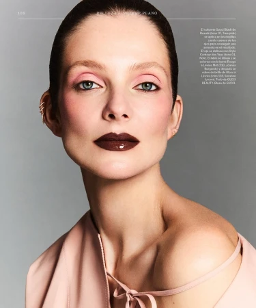 Beauty shot of a woman with pink eye makeup and dark glossy lipstick, posing in front of a gray background. She wears a pink top.