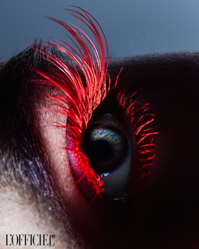Close-up fashion beauty shot of a blue eye with bright red mascara on the lashes against a dark gray background, magazine logo visible.
