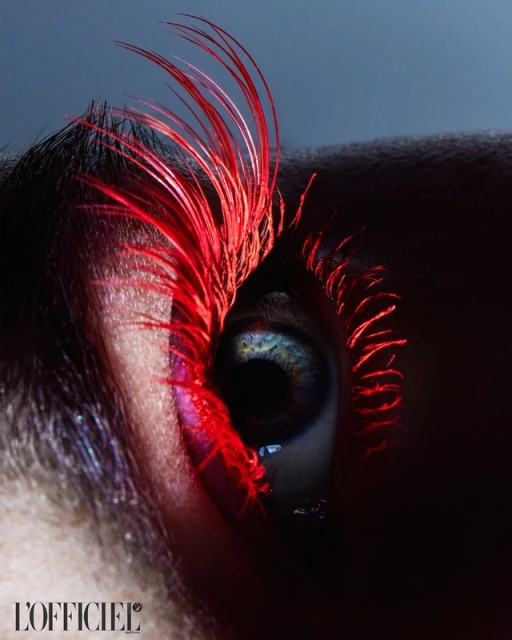 Close-up fashion beauty shot of a blue eye with bright red mascara on the lashes against a dark gray background, magazine logo visible.
