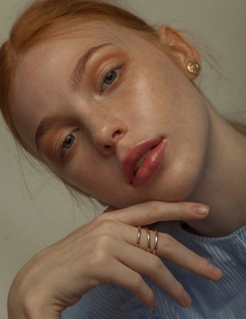 Close-up portrait of a red-haired woman with fair skin, wearing gold rings and a blue striped top, posing indoors with soft lighting.