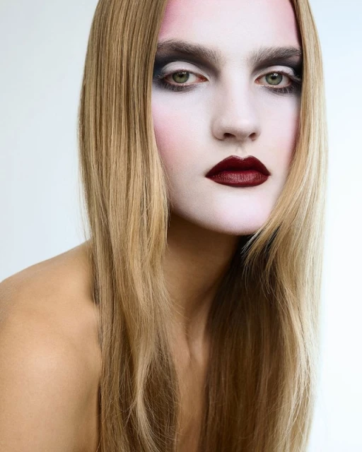 Close-up studio portrait of a woman with pale skin, blonde hair, heavy dark eye makeup, and dark red lipstick against a white backdrop.