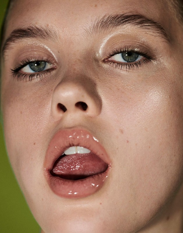Close-up portrait of an adult woman with shiny skin and wet pink lips, set against a bright green background, showcasing a beauty or fashion aesthetic.