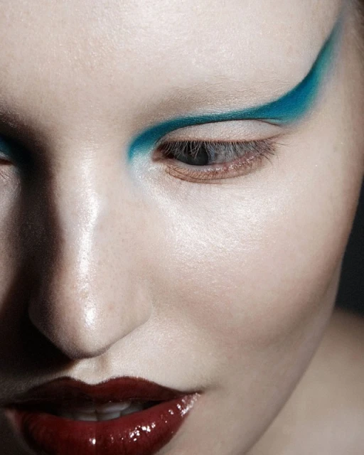 Close-up of a woman's face featuring dramatic turquoise eyeshadow on her upper eyelid and glossy, dark red lips against a dark background.