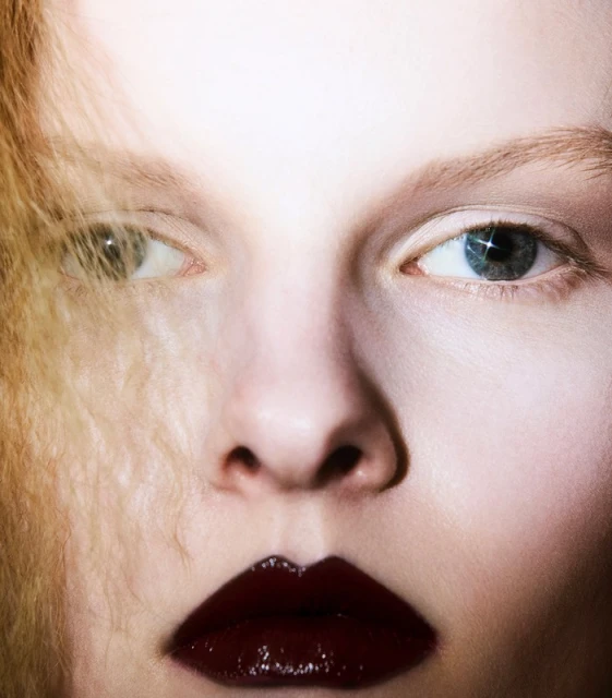 Close-up portrait of a fair-skinned woman with dark red lipstick. Golden hair strands are visible. The eyes are different colors.