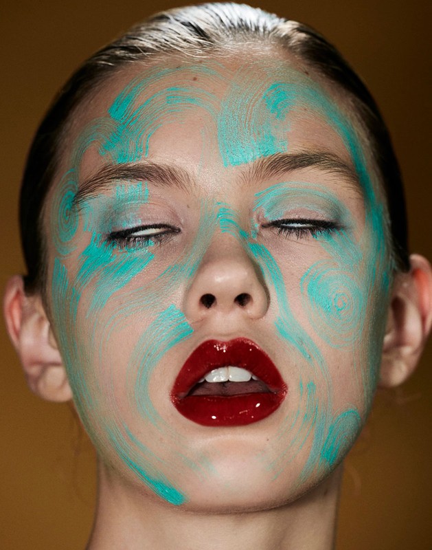 Close-up of a woman's face with abstract turquoise face paint and glossy red lips against a mustard yellow background in a beauty shot.