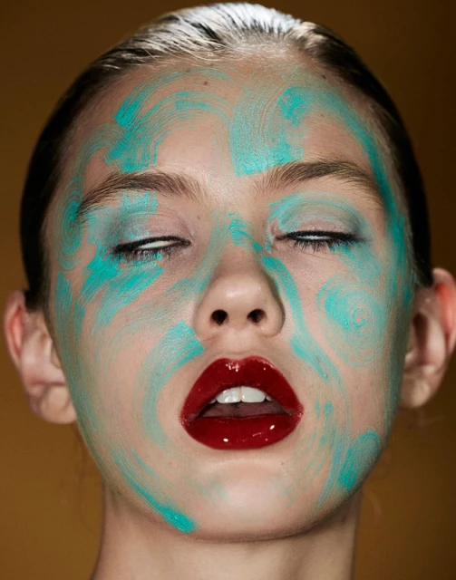 Close-up of a woman's face with abstract turquoise face paint and glossy red lips against a mustard yellow background in a beauty shot.