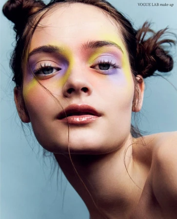 Close-up portrait of a woman with vibrant yellow and purple avant-garde eye makeup and double hair buns against a blue background.
