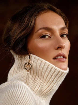 Close-up portrait of a woman wearing a cream ribbed turtleneck sweater and large gold earrings with makeup by David Razzano