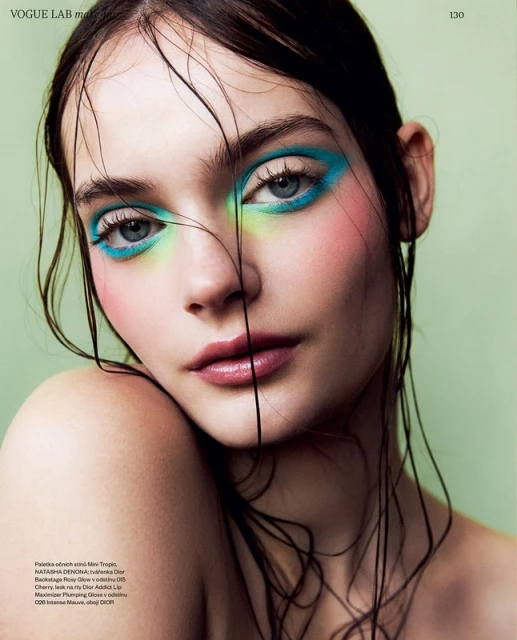Close-up of a woman with wet hair and colorful makeup. The model has green, blue, and pink eye shadow with glossy lips.