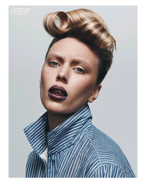 A portrait of a woman with retro blonde hair, shiny dark lipstick, wearing a striped shirt, against a gray background in a beauty editorial.