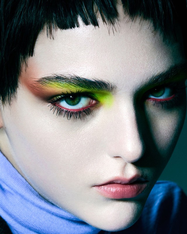 Close-up portrait of a woman with green eyes, neon eye makeup, and dark, blunt-cut bangs looking directly at the camera in a studio setting.