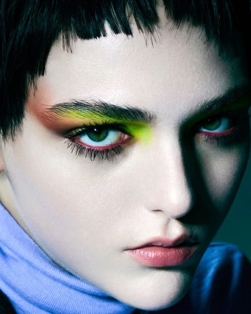 Close-up portrait of a woman with green eyes, neon eye makeup, and dark, blunt-cut bangs looking directly at the camera in a studio setting.