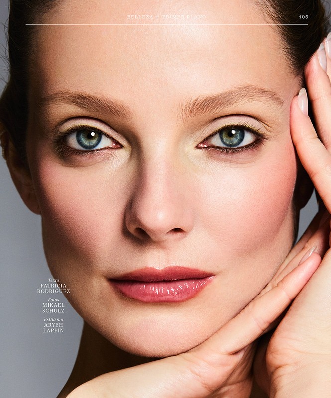 Close-up portrait of a woman with blue eyes, subtle makeup, and hands touching her face against a neutral background