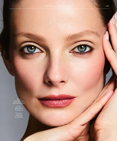 Close-up portrait of a woman with blue eyes, subtle makeup, and hands touching her face against a neutral background