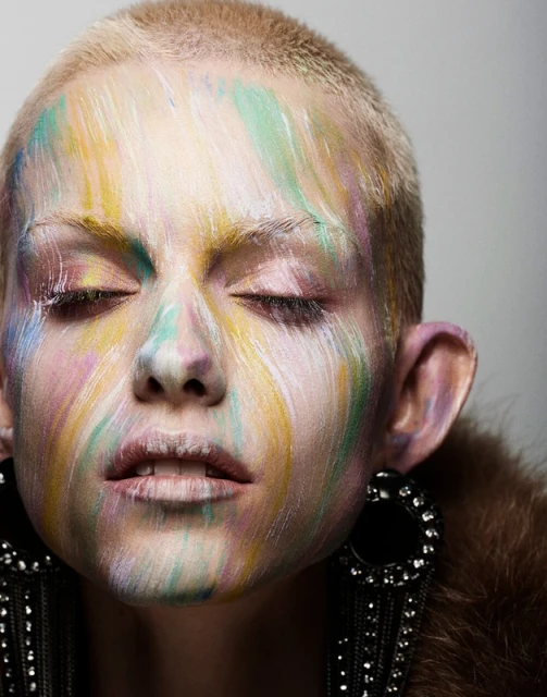 Woman with short blonde hair and eyes closed, face adorned with streaks of pastel paint in pink, yellow, blue, and white hues.