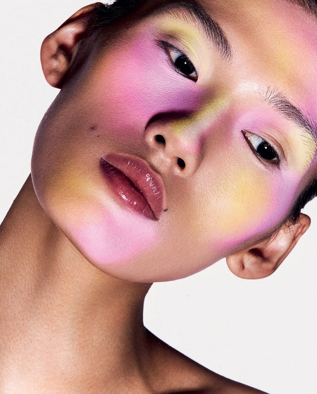 Close-up portrait of a person with multicolored pastel face makeup in shades of pink, yellow, and purple against a white background.