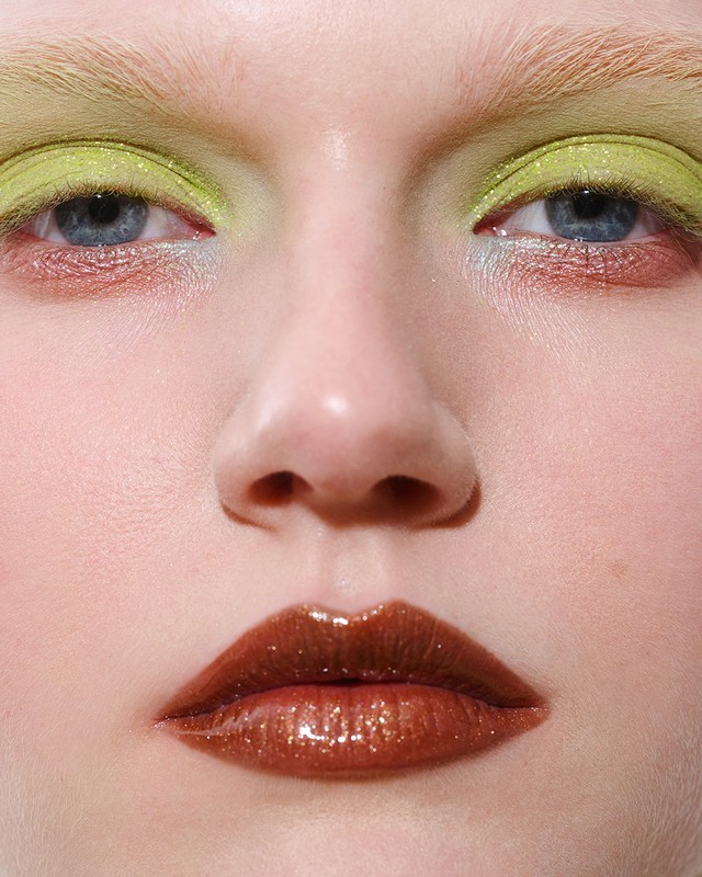 Close-up of a woman's face with bright green eyeshadow and glossy bronze lipstick, showcasing makeup artistry and beauty details.