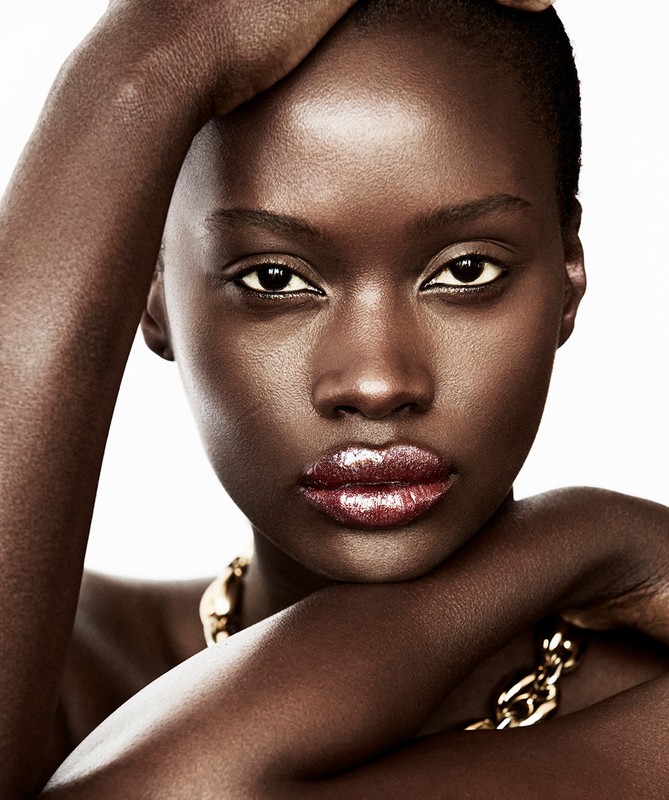 Close-up portrait of a striking dark-skinned woman with glossy lips and a gold chain necklace, set against a bright white background.
