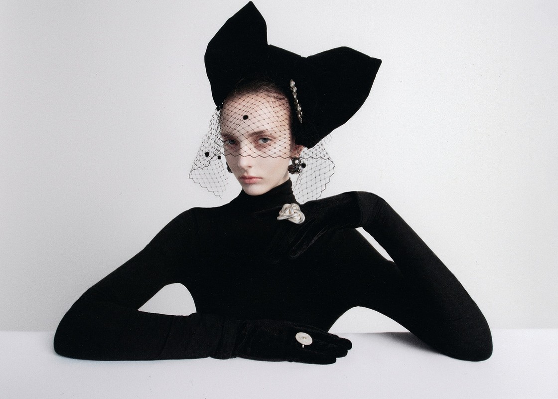 A model poses wearing a large black bow headpiece and black gloves against a white background in a studio fashion photograph.