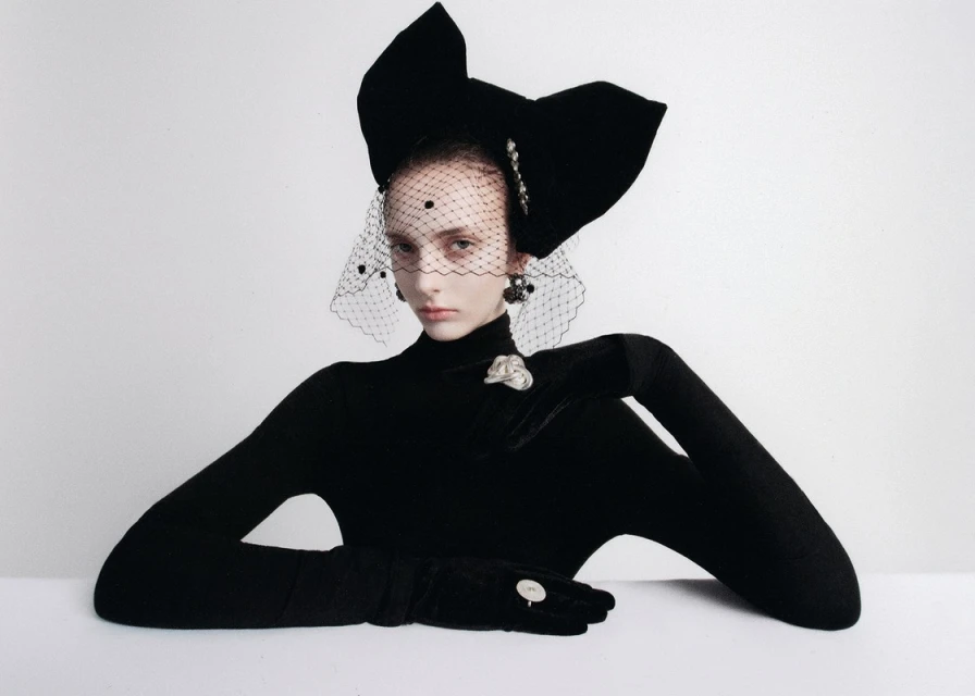 A model poses wearing a large black bow headpiece and black gloves against a white background in a studio fashion photograph.