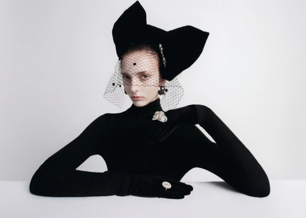 A model poses wearing a large black bow headpiece and black gloves against a white background in a studio fashion photograph.