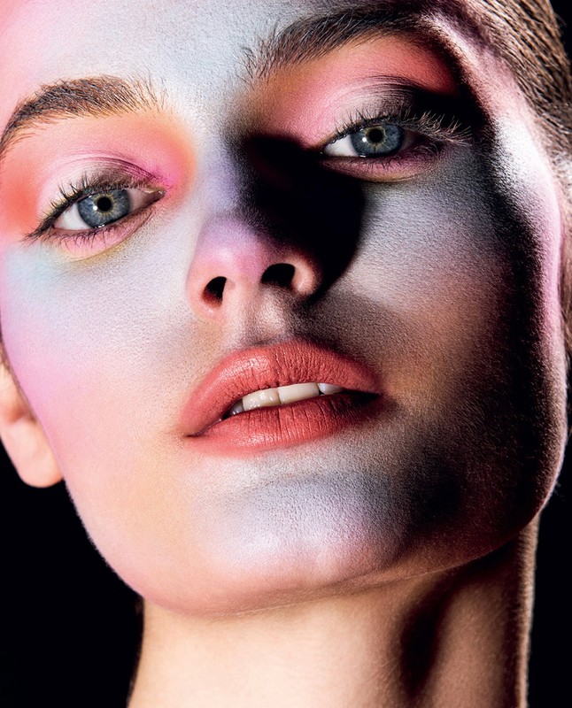 Close-up portrait of a woman with blue eyes, light skin, and colorful face paint in shades of pink, orange, and blue against a black background.