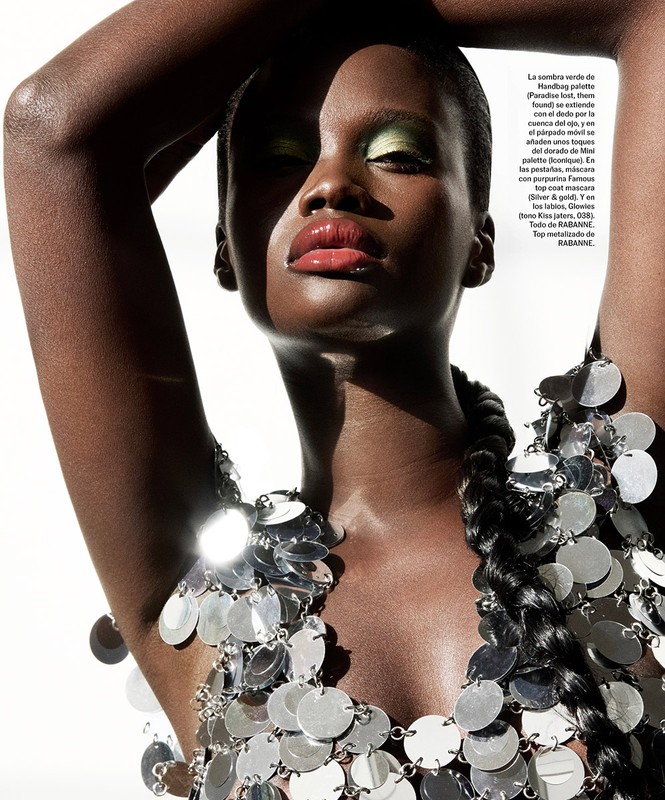 Fashion shot of a Black female model wearing green eyeshadow and a shiny silver disc top with hands above her head in an indoor setting.