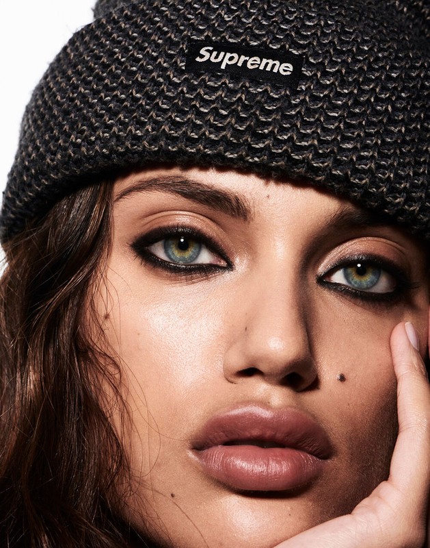 Close-up portrait of a woman with hazel eyes and dark hair wearing a gray Supreme beanie against a white background.