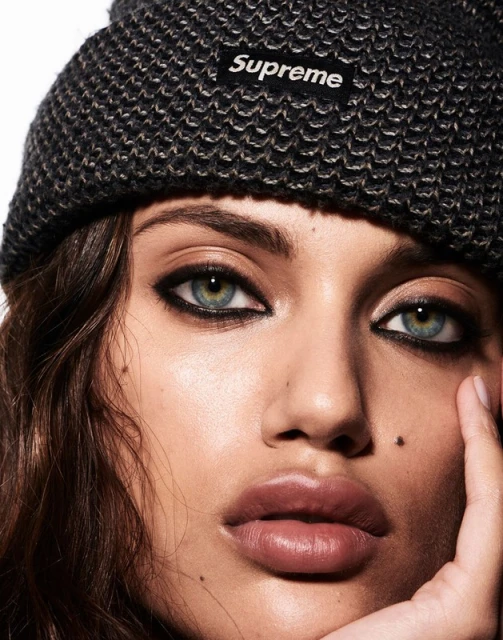 Close-up portrait of a woman with hazel eyes and dark hair wearing a gray Supreme beanie against a white background.