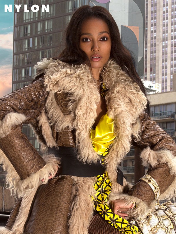 A fashion portrait features a woman with long brown hair wearing a patterned brown and cream fur-trimmed jacket with city buildings behind.