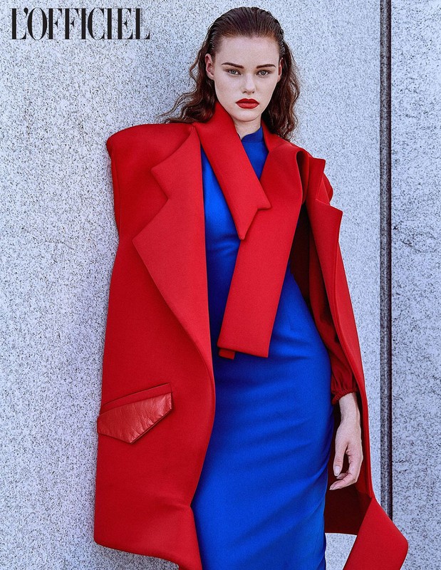 Fashion shot of a female model wearing a bold red coat and blue dress against a concrete wall with the L'Officiel magazine logo.
