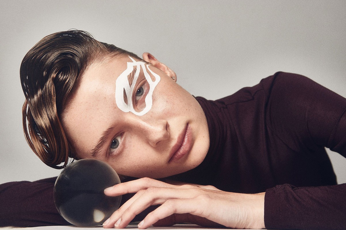 Woman with stylized eye makeup and slicked-back hair poses with a crystal ball in a portrait with a white background and neutral lighting.