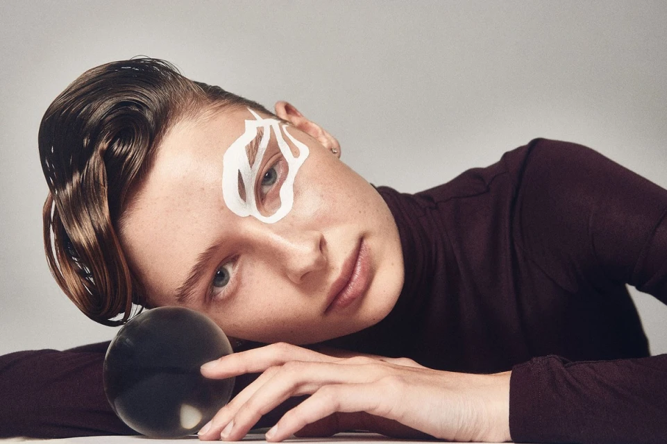 Woman with stylized eye makeup and slicked-back hair poses with a crystal ball in a portrait with a white background and neutral lighting.