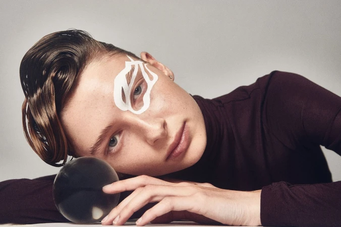 Woman with stylized eye makeup and slicked-back hair poses with a crystal ball in a portrait with a white background and neutral lighting.